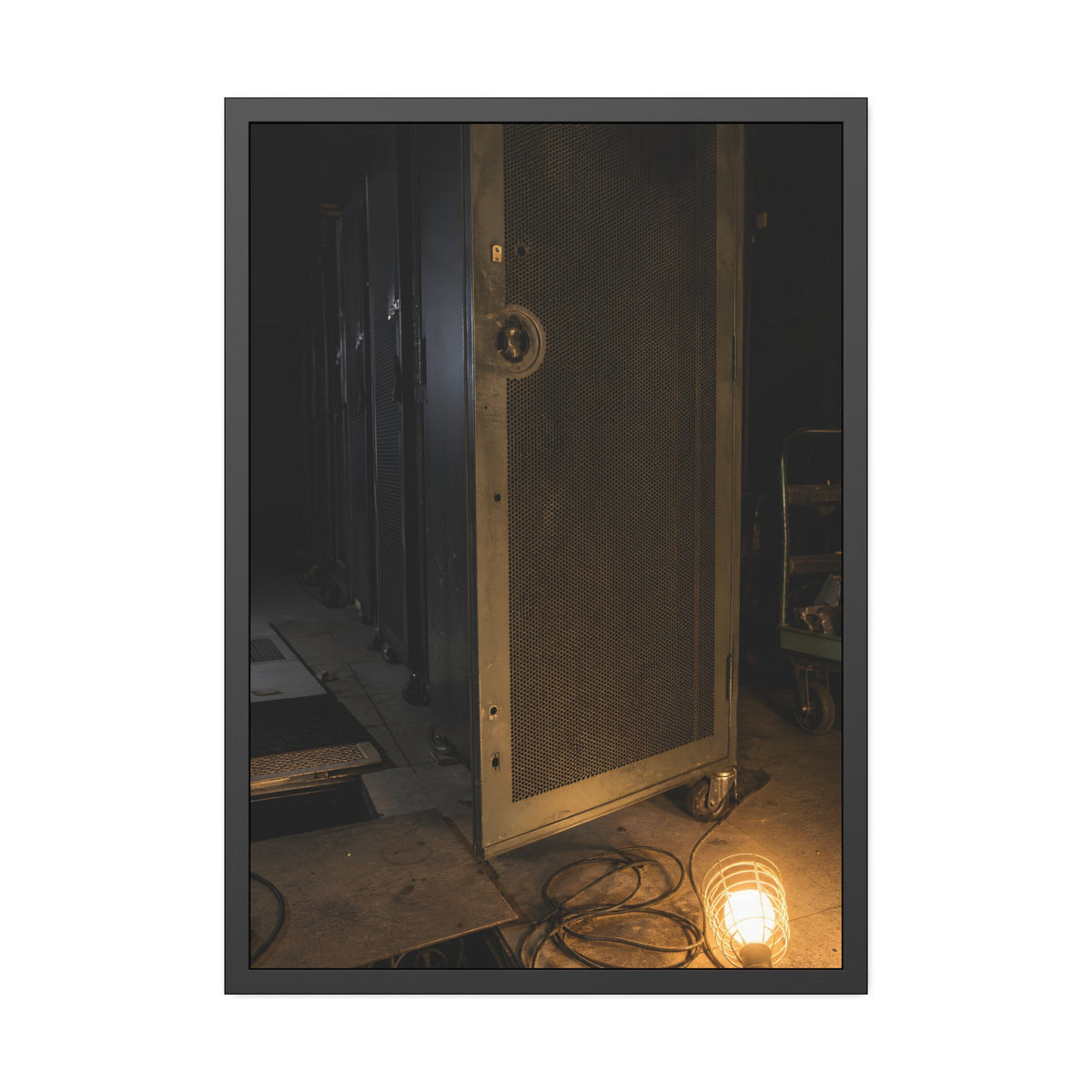 A perforated server rack door in a dark aisle lit by a floor work lamp. The Cold Aisle by Garabaggio, framed fine art print, 28″ x 40″.