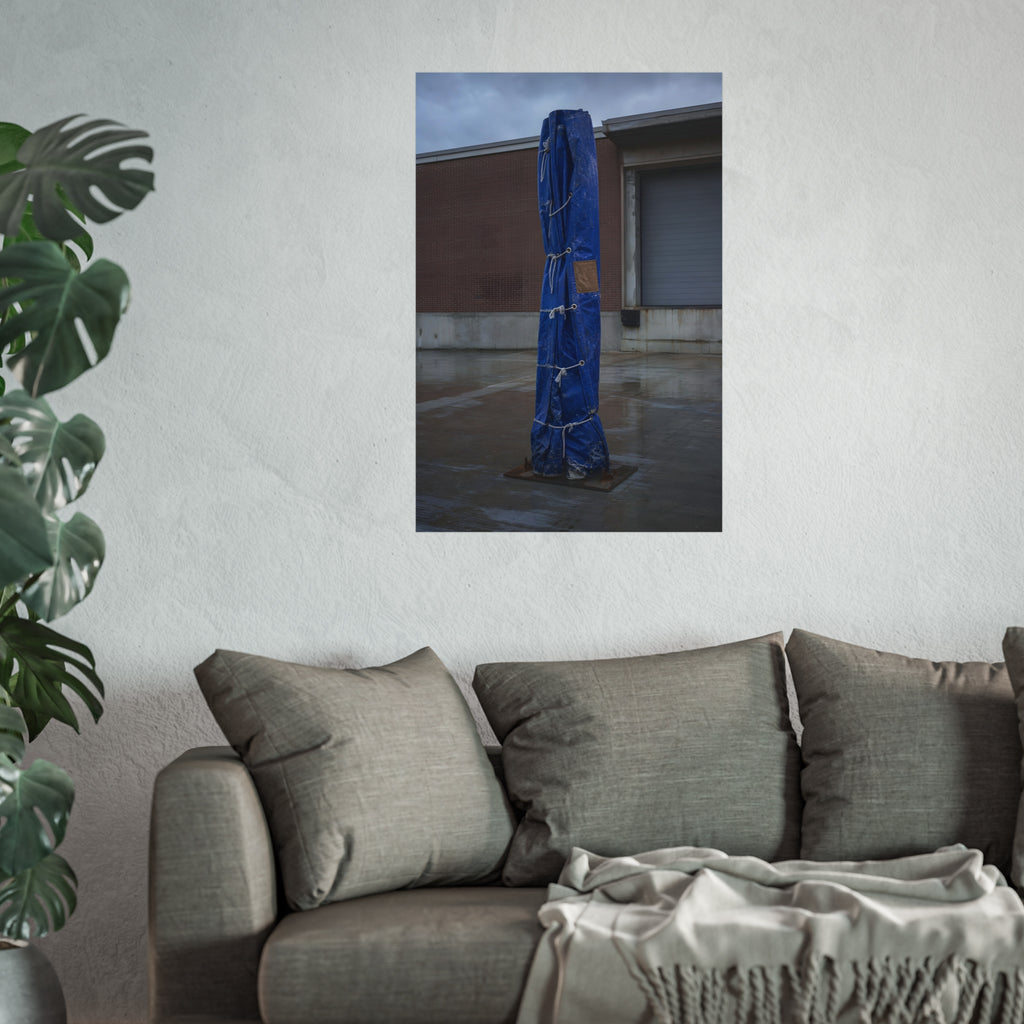 A tall blue tarp bundle roped upright on a wet lot by a loading dock. Folded Standard at the Loading Bay by Garabaggio, fine art print hanging in a room.