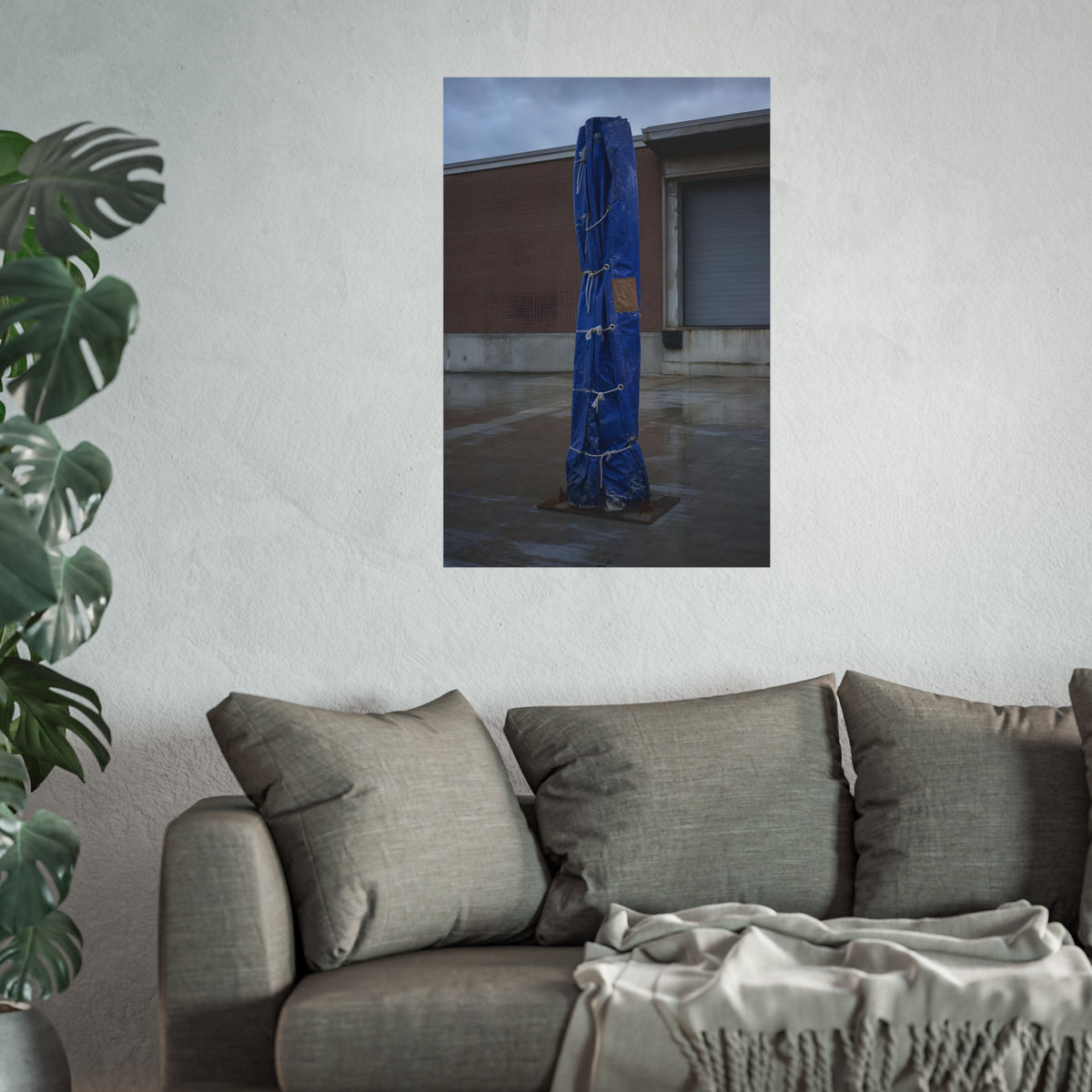 A tall blue tarp bundle roped upright on a wet lot by a loading dock. Folded Standard at the Loading Bay by Garabaggio, fine art print hanging in a room.