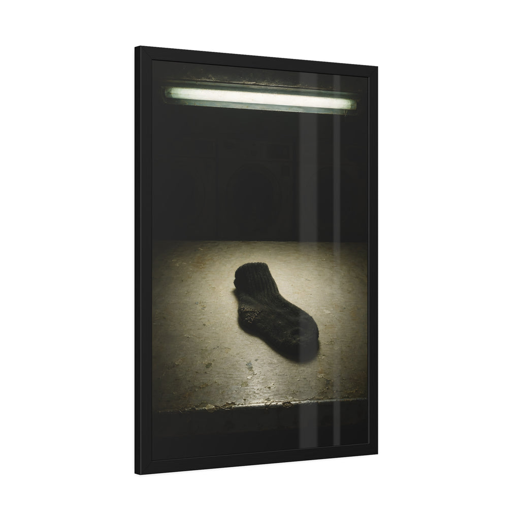 A single dark sock on a laundromat floor before dim washing machines. The Orphaned Sock by Garabaggio, framed fine art print hanging in a room.