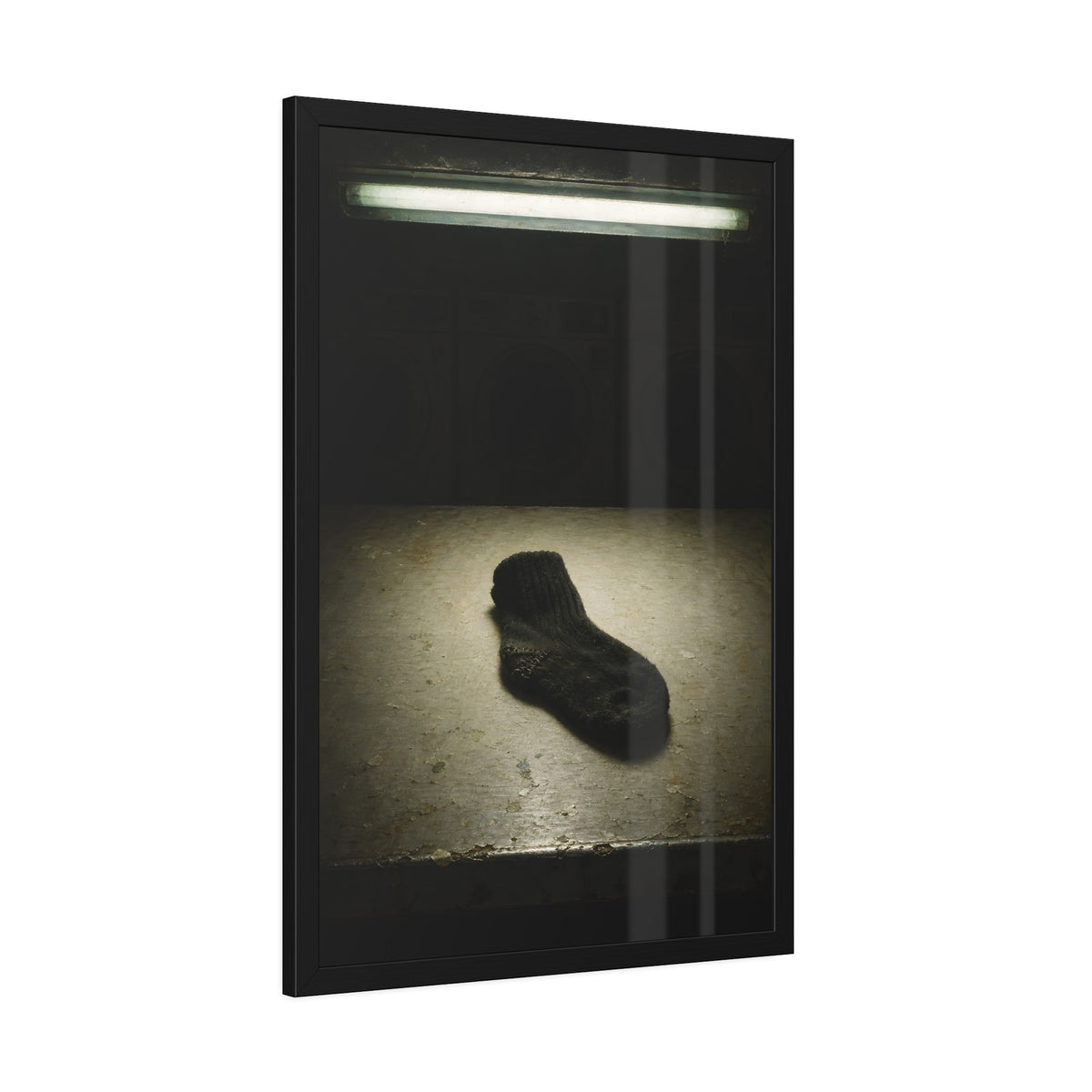 A single dark sock on a laundromat floor before dim washing machines. The Orphaned Sock by Garabaggio, framed fine art print hanging in a room.