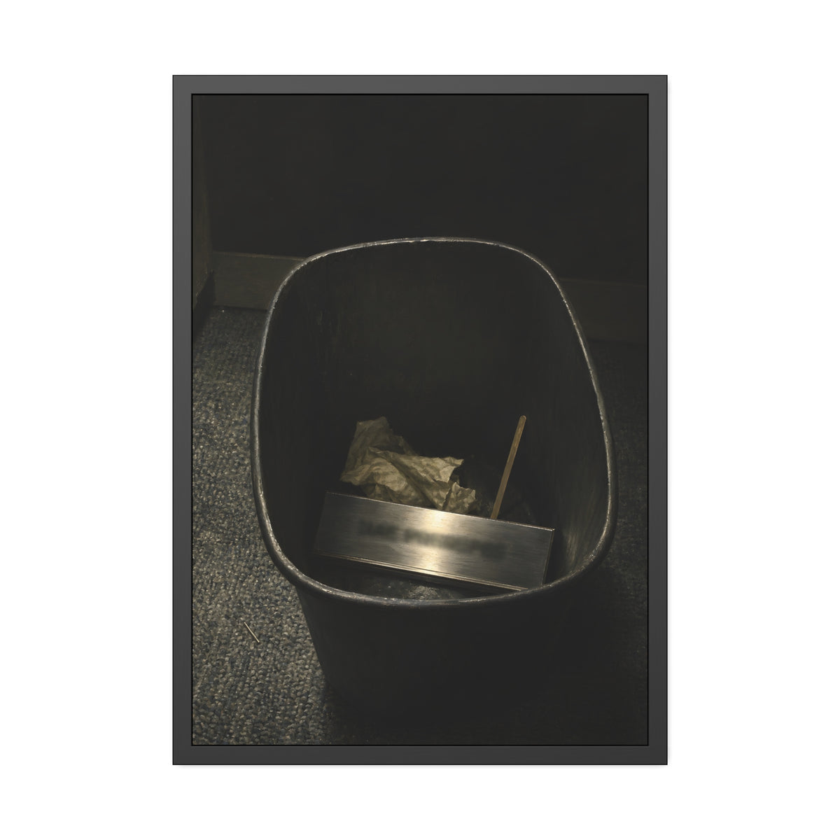 A metal nameplate lying in a black office waste bin on grey carpet. The Nameplate in the Waste Bin by Garabaggio, framed fine art print, 28″ x 40″.