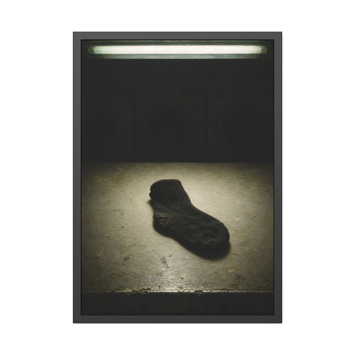 A single dark sock on a laundromat floor before dim washing machines. The Orphaned Sock by Garabaggio, framed fine art print, 28″ x 40″.