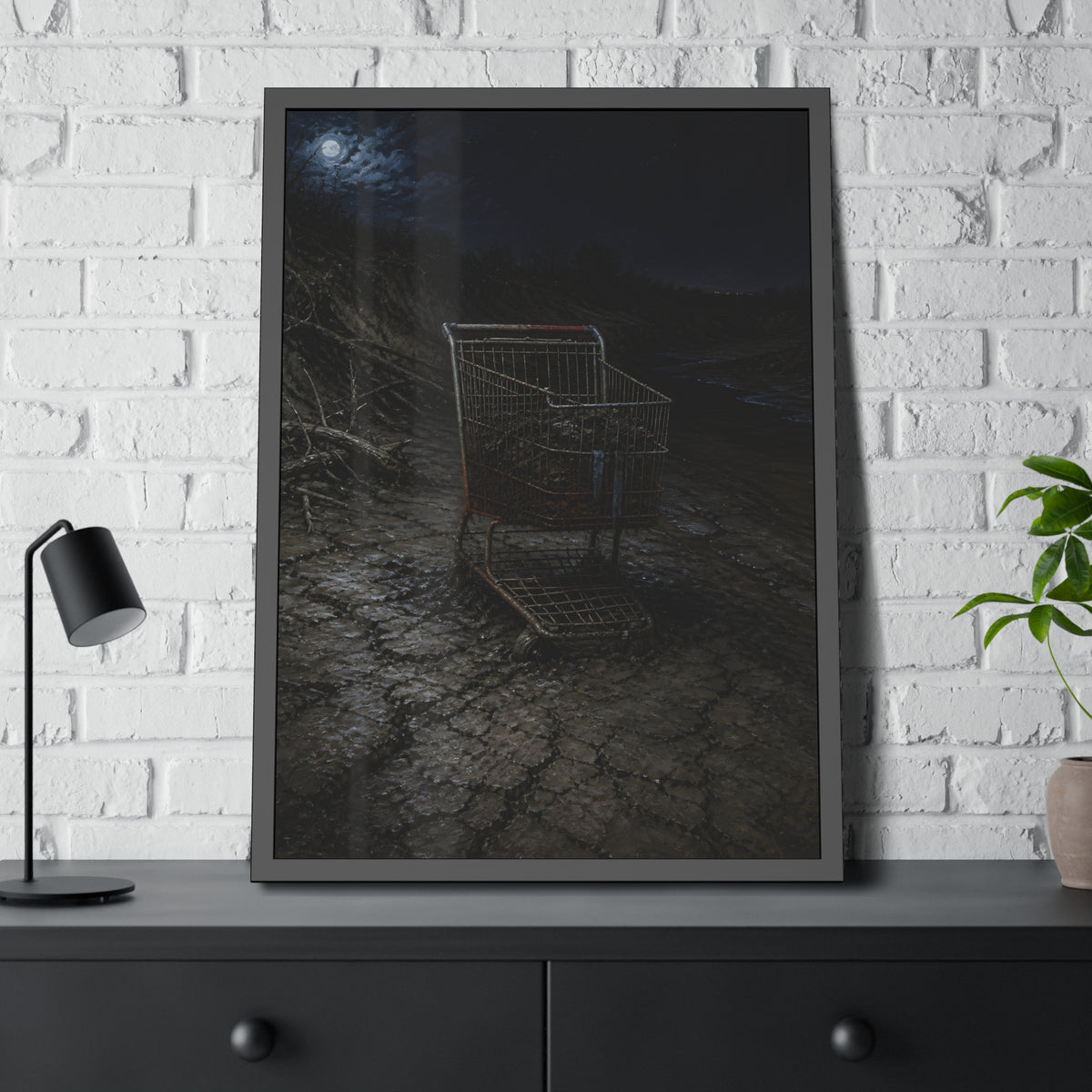 A rusted shopping cart sunk in cracked dried mud under a full moon. The Silting by Garabaggio, framed fine art print hanging in a room.