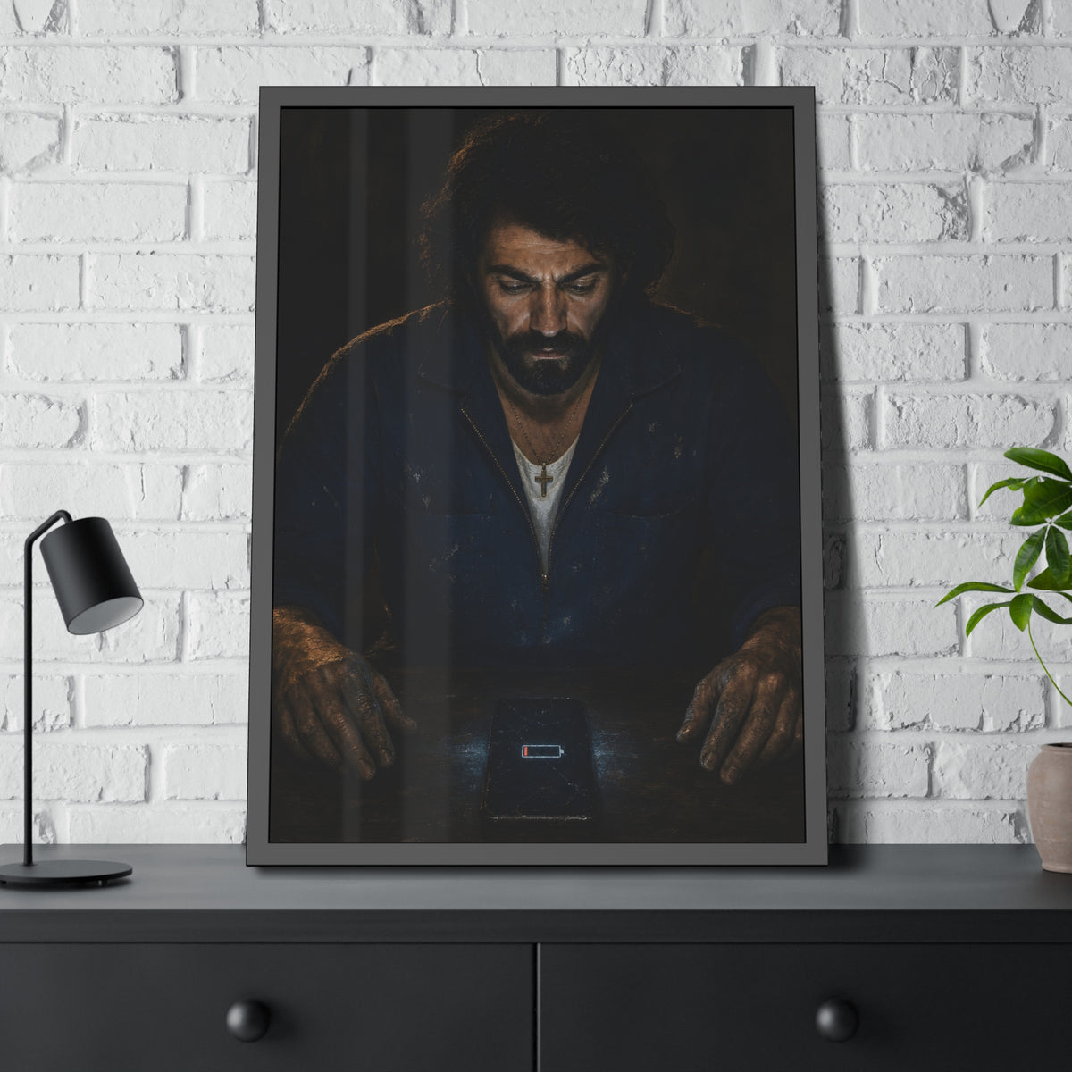 A man in a work jacket looks down at a phone showing a red low battery icon. The Calling at One Percent by Garabaggio, framed fine art print hanging in a room.
