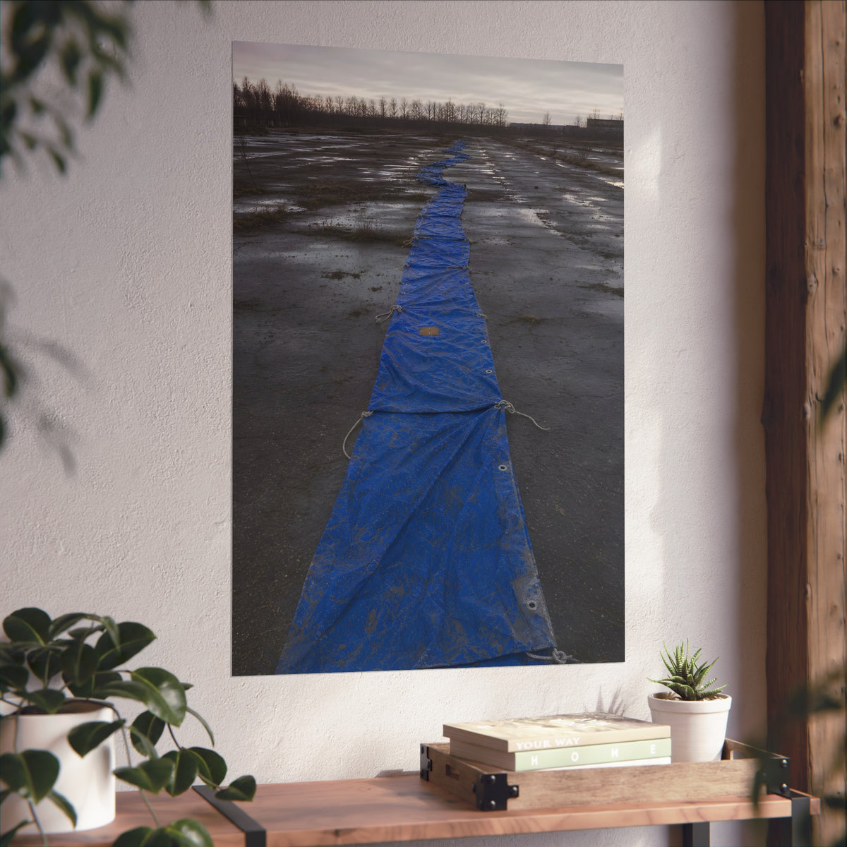 A blue tarp laid in a long strip down a wet cracked lot under grey sky. The Long Ground Procession by Garabaggio, fine art print hanging in a room.