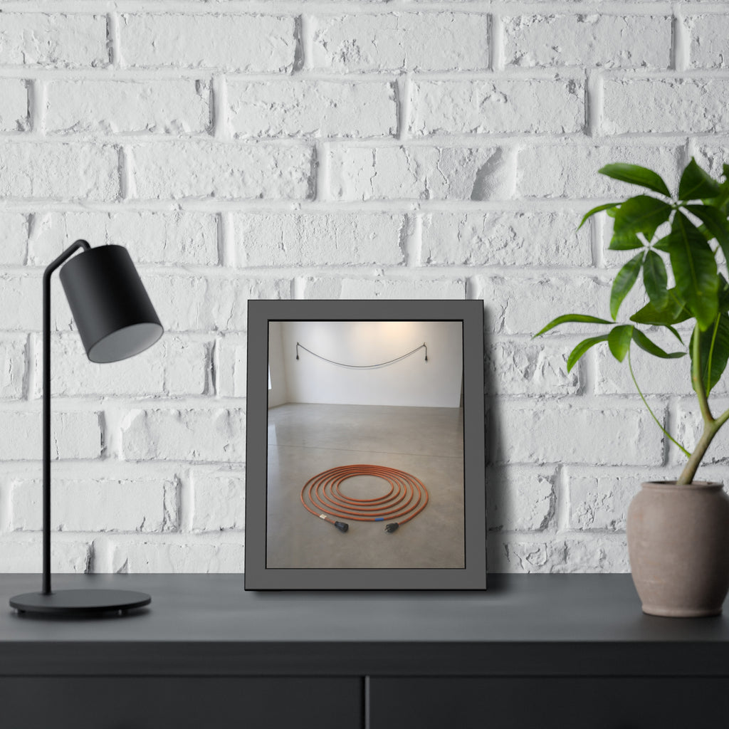 An orange extension cord coiled in a spiral on a bare gallery floor. The Floor Coil by Garabaggio, framed fine art print hanging in a room.
