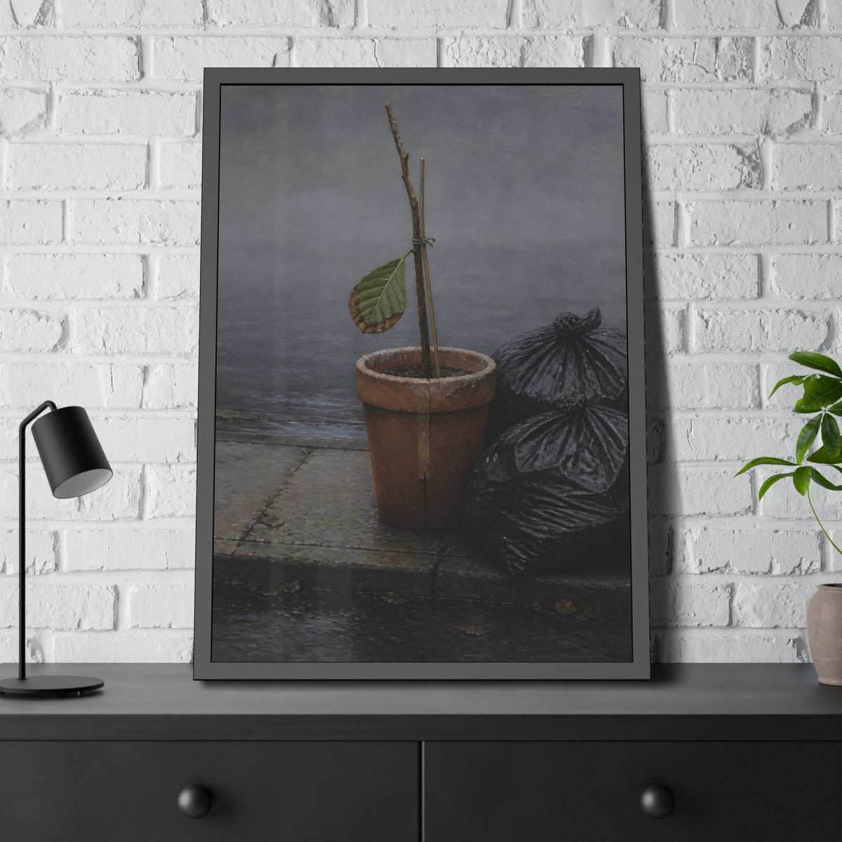 A potted dead twig with one leaf beside black garbage bags on a wet curb. The Curb on Tuesday by Garabaggio, framed fine art print hanging in a room.