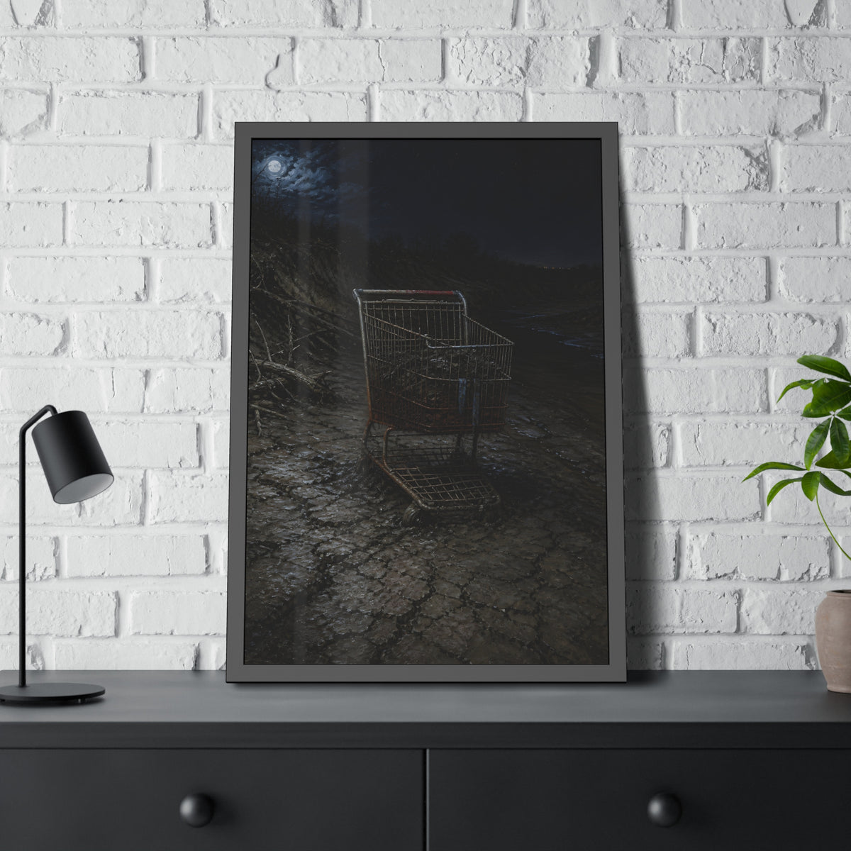 A rusted shopping cart sunk in cracked dried mud under a full moon. The Silting by Garabaggio, framed fine art print hanging in a room.