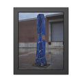 A tall blue tarp bundle roped upright on a wet lot by a loading dock. Folded Standard at the Loading Bay by Garabaggio, framed fine art print, 11″ x 14″.