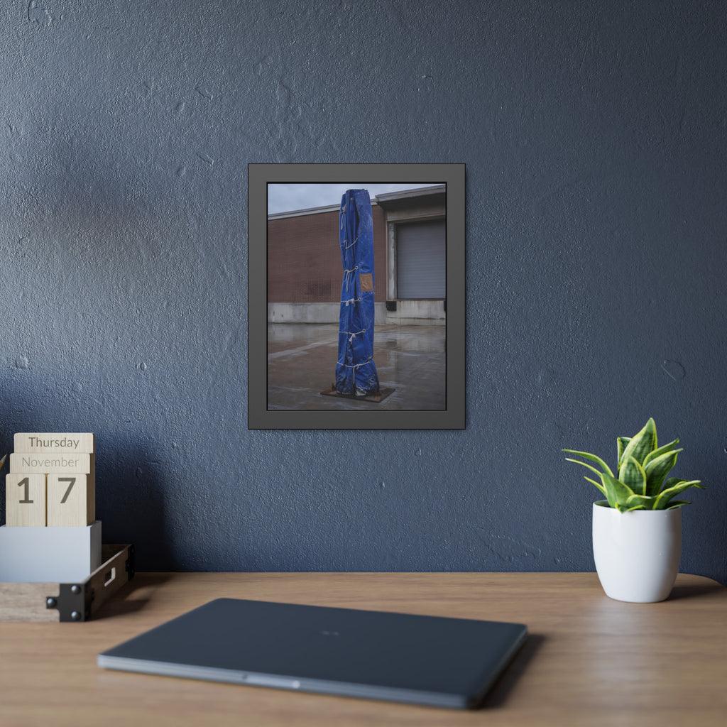 A tall blue tarp bundle roped upright on a wet lot by a loading dock. Folded Standard at the Loading Bay by Garabaggio, framed fine art print hanging in a room.