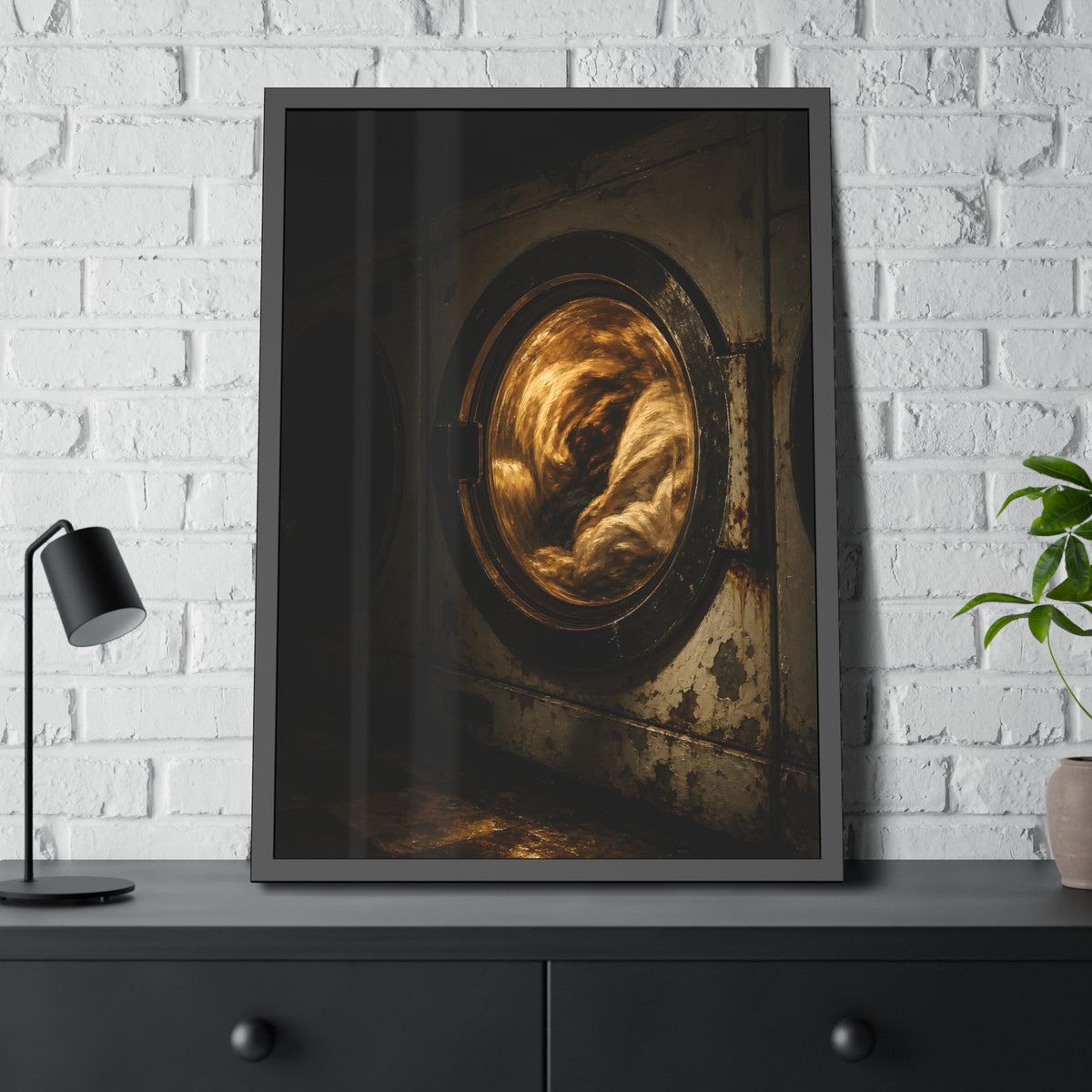 A washer drum tumbling laundry lit amber behind its glass door. The First Spin by Garabaggio, framed fine art print hanging in a room.