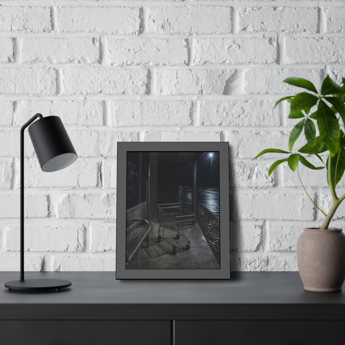 A shopping cart parked inside a bus shelter under a street lamp at night. The Rest at the Bus Shelter by Garabaggio, framed fine art print hanging in a room.