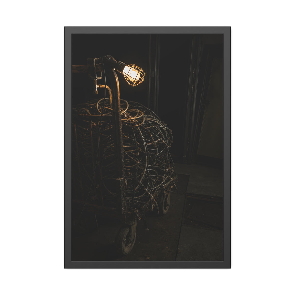 A tangled ball of cables on a hand cart under a caged work lamp. The Cable Harvest by Garabaggio, framed fine art print, 24″ x 36″.