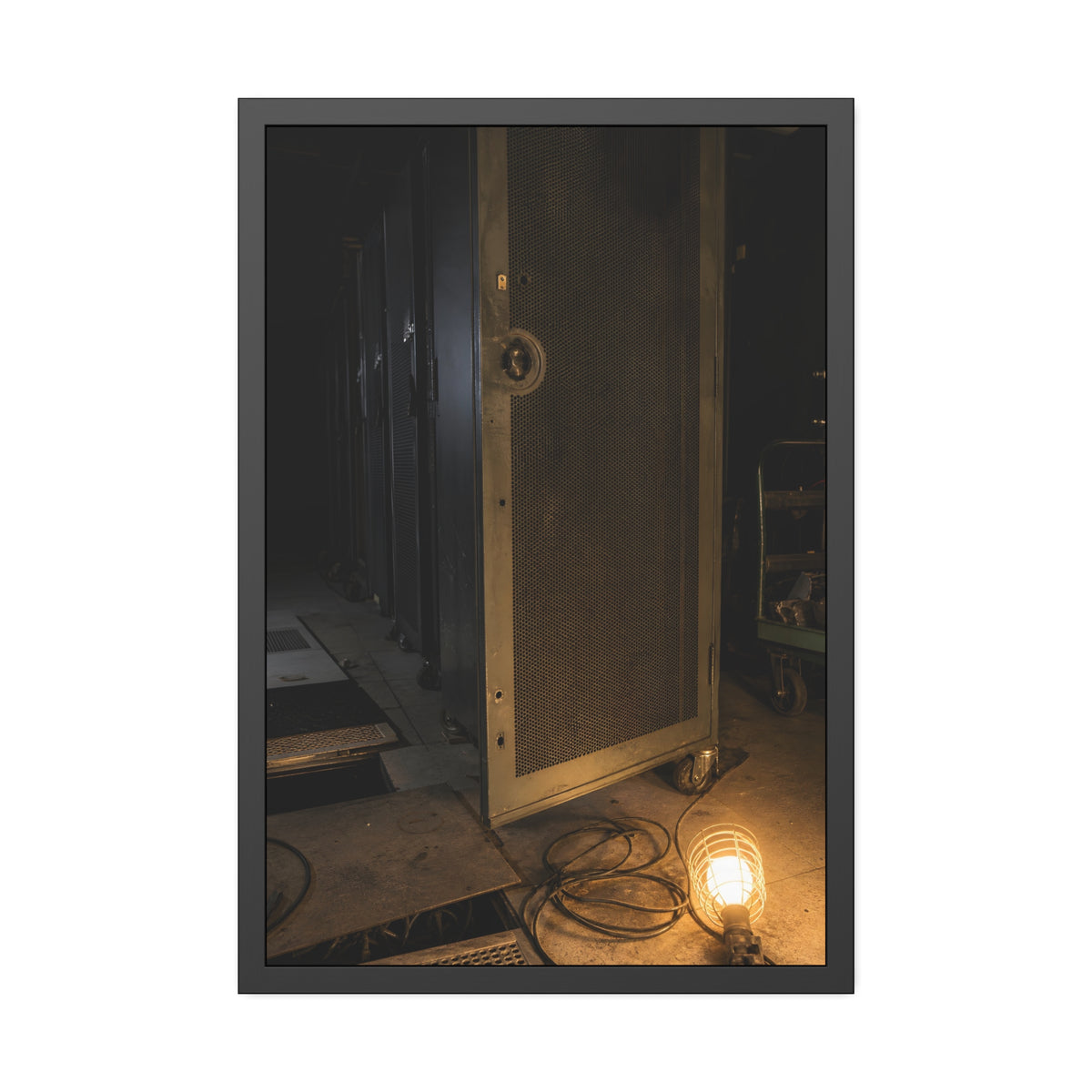 A perforated server rack door in a dark aisle lit by a floor work lamp. The Cold Aisle by Garabaggio, framed fine art print, 24″ x 36″.