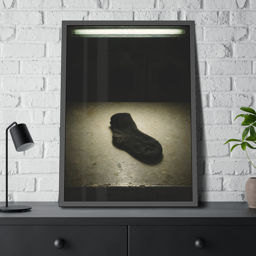 A single dark sock on a laundromat floor before dim washing machines. The Orphaned Sock by Garabaggio, framed fine art print hanging in a room.
