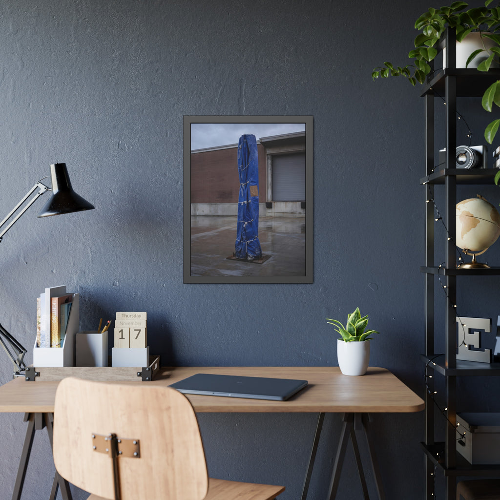 A tall blue tarp bundle roped upright on a wet lot by a loading dock. Folded Standard at the Loading Bay by Garabaggio, framed fine art print hanging in a room.