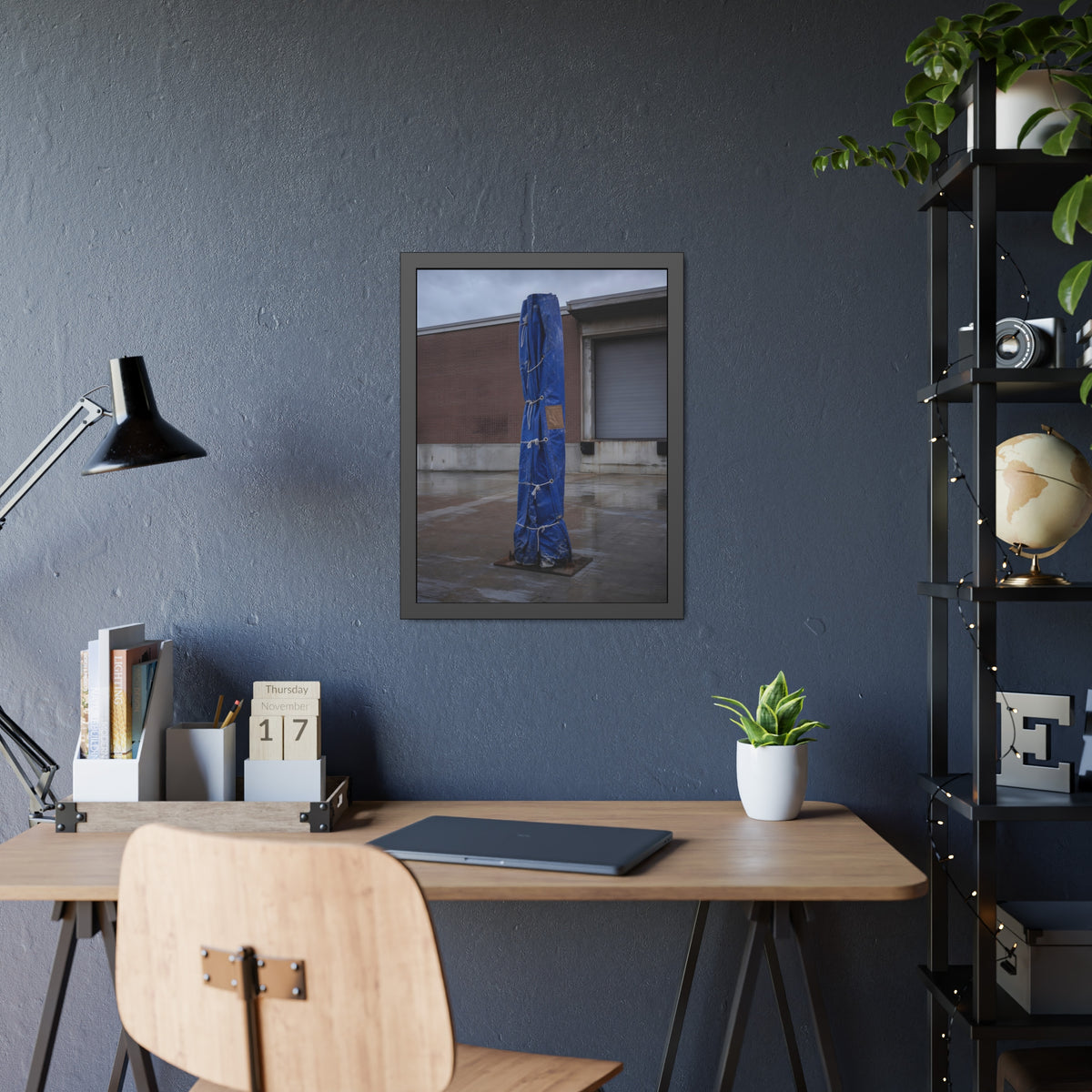 A tall blue tarp bundle roped upright on a wet lot by a loading dock. Folded Standard at the Loading Bay by Garabaggio, framed fine art print hanging in a room.