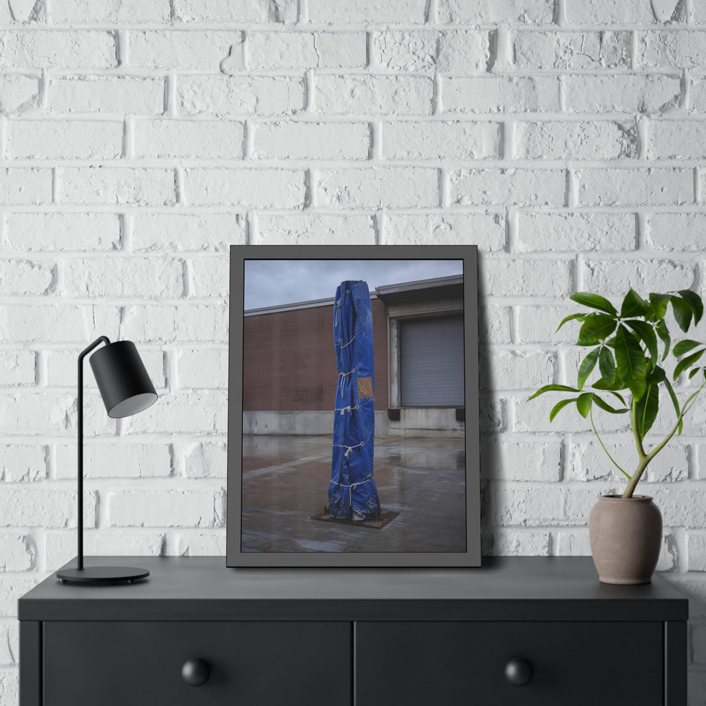 A tall blue tarp bundle roped upright on a wet lot by a loading dock. Folded Standard at the Loading Bay by Garabaggio, framed fine art print hanging in a room.