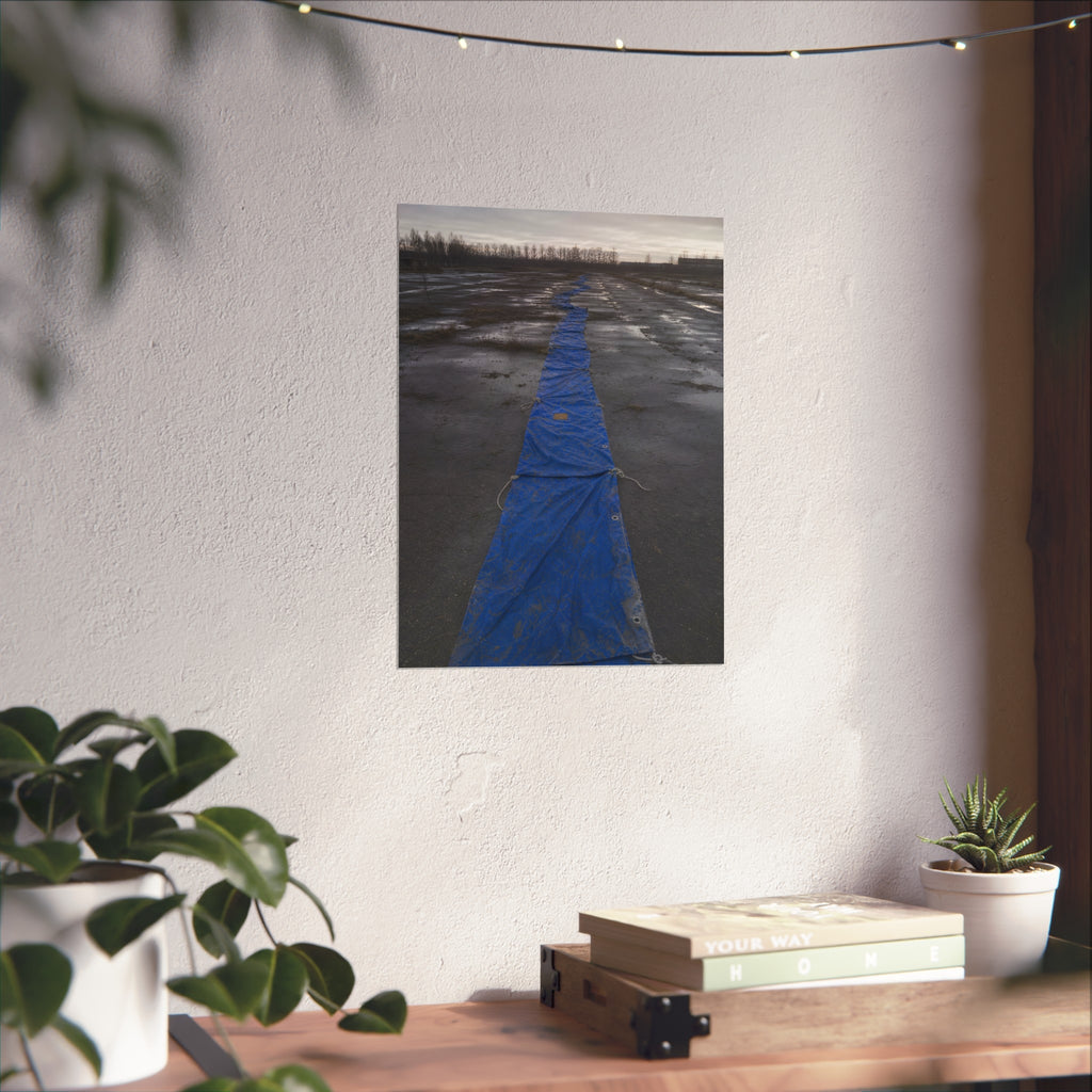 A blue tarp laid in a long strip down a wet cracked lot under grey sky. The Long Ground Procession by Garabaggio, fine art print hanging in a room.