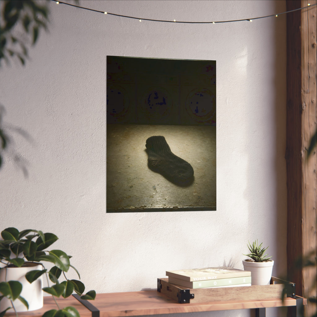 A single dark sock on a laundromat floor before dim washing machines. The Orphaned Sock by Garabaggio, fine art print hanging in a room.