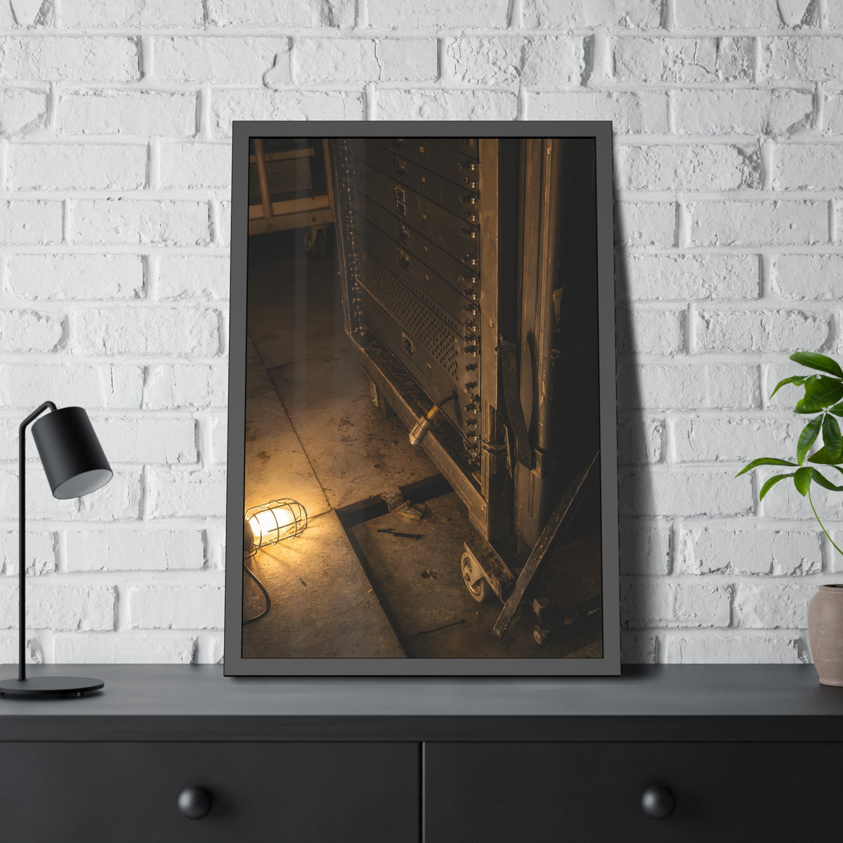 A server rack on a concrete floor beside a caged work light. The Silence at Cutover by Garabaggio, framed fine art print hanging in a room.