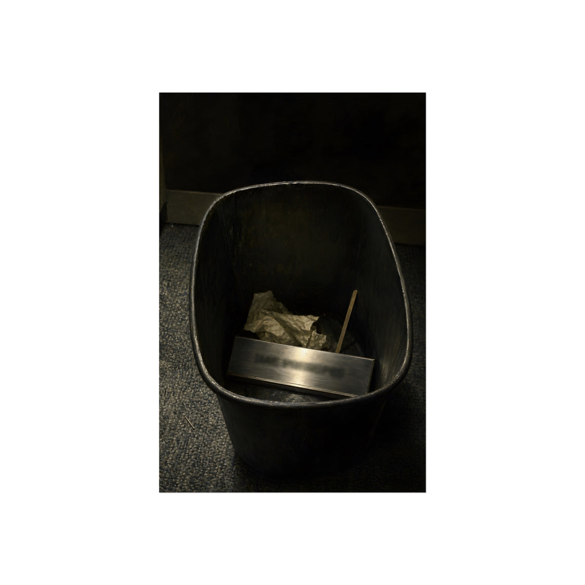A metal nameplate lying in a black office waste bin on grey carpet. The Nameplate in the Waste Bin by Garabaggio, fine art print, 24″ x 36″.