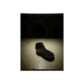 A single dark sock on a laundromat floor before dim washing machines. The Orphaned Sock by Garabaggio, fine art print, 12″ x 16″.