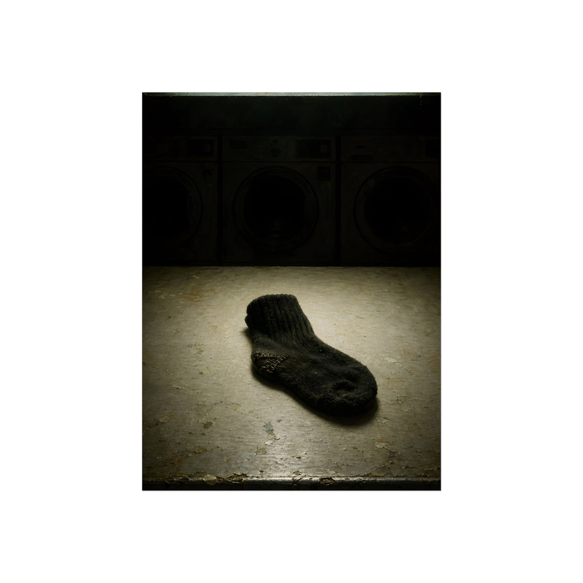 A single dark sock on a laundromat floor before dim washing machines. The Orphaned Sock by Garabaggio, fine art print, 12″ x 16″.