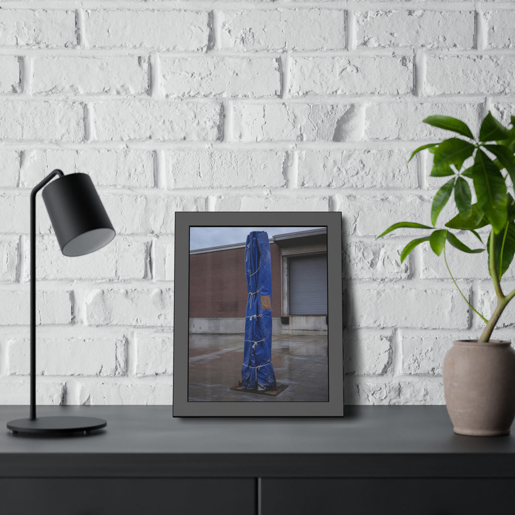 A tall blue tarp bundle roped upright on a wet lot by a loading dock. Folded Standard at the Loading Bay by Garabaggio, framed fine art print hanging in a room.