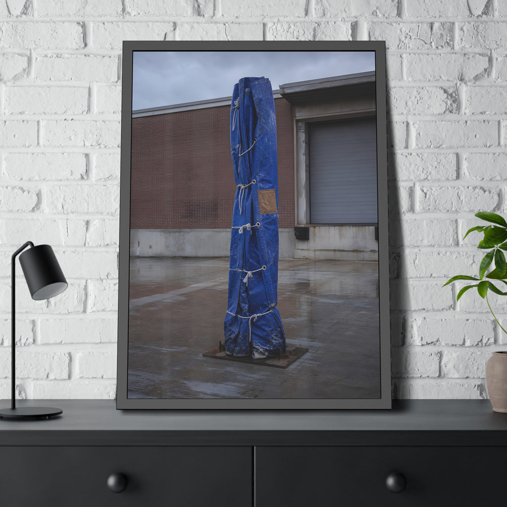 A tall blue tarp bundle roped upright on a wet lot by a loading dock. Folded Standard at the Loading Bay by Garabaggio, framed fine art print hanging in a room.
