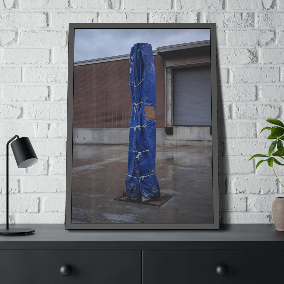 A tall blue tarp bundle roped upright on a wet lot by a loading dock. Folded Standard at the Loading Bay by Garabaggio, framed fine art print hanging in a room.