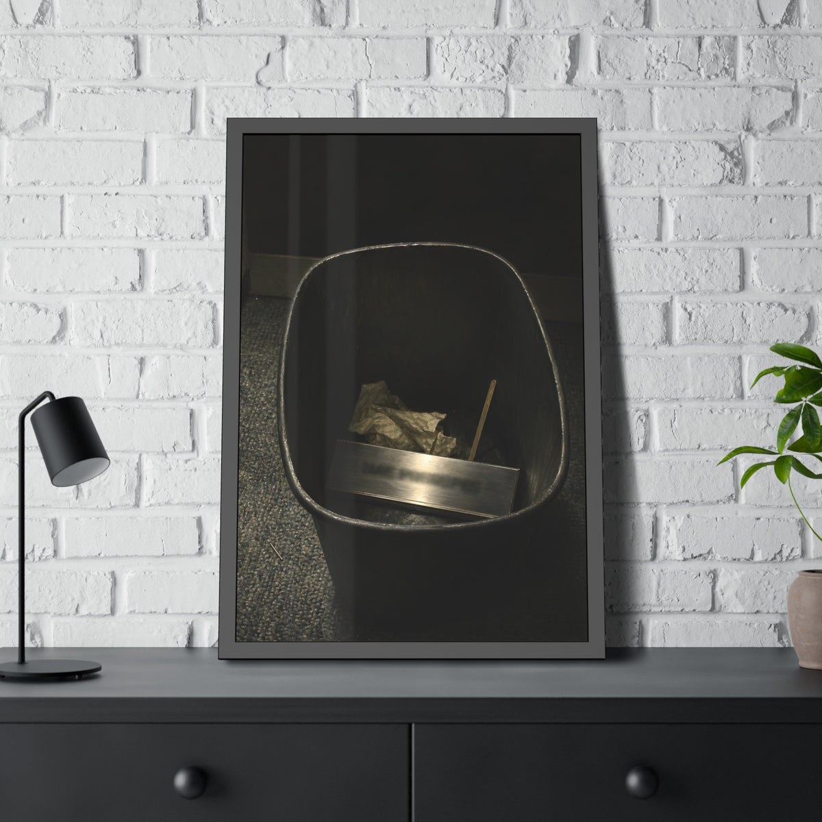 A metal nameplate lying in a black office waste bin on grey carpet. The Nameplate in the Waste Bin by Garabaggio, framed fine art print hanging in a room.