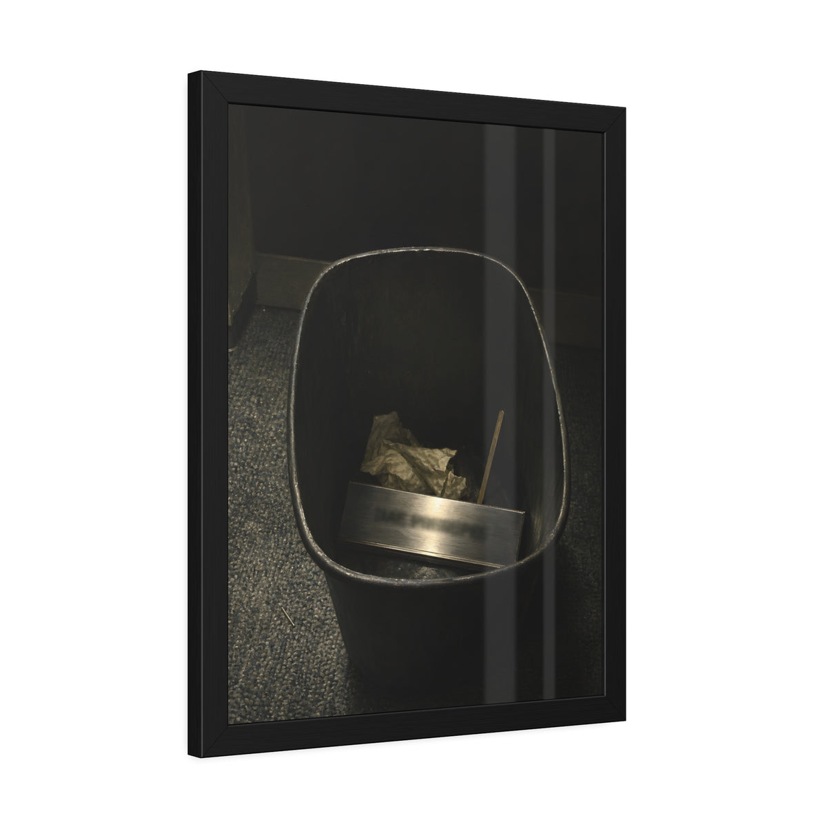 A metal nameplate lying in a black office waste bin on grey carpet. The Nameplate in the Waste Bin by Garabaggio, framed fine art print hanging in a room.