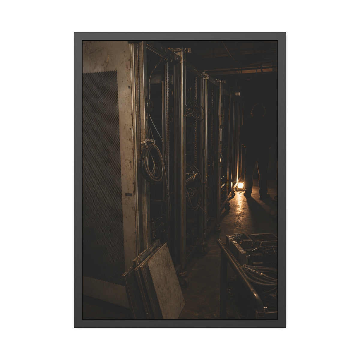 Open server racks in a dark room, a work lamp and a figure at the end. Garabaggio at the End of the Row by Garabaggio, framed fine art print, 28″ x 40″.