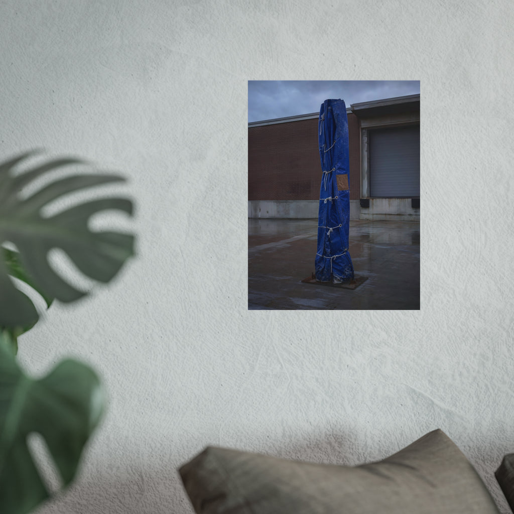 A tall blue tarp bundle roped upright on a wet lot by a loading dock. Folded Standard at the Loading Bay by Garabaggio, fine art print hanging in a room.