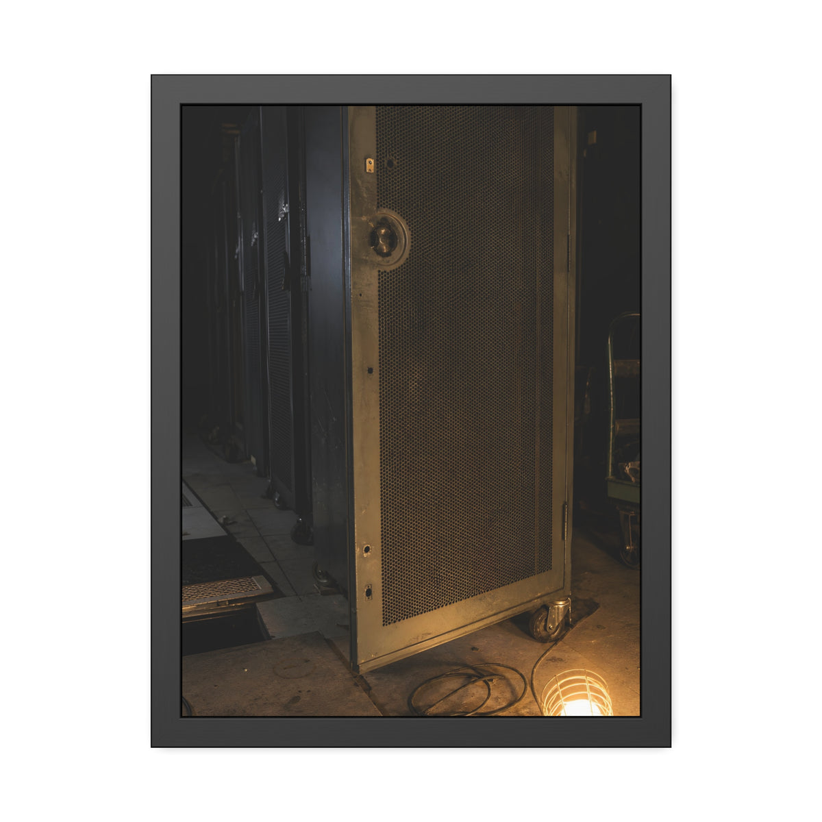A perforated server rack door in a dark aisle lit by a floor work lamp. The Cold Aisle by Garabaggio, framed fine art print, 18″ x 24″.