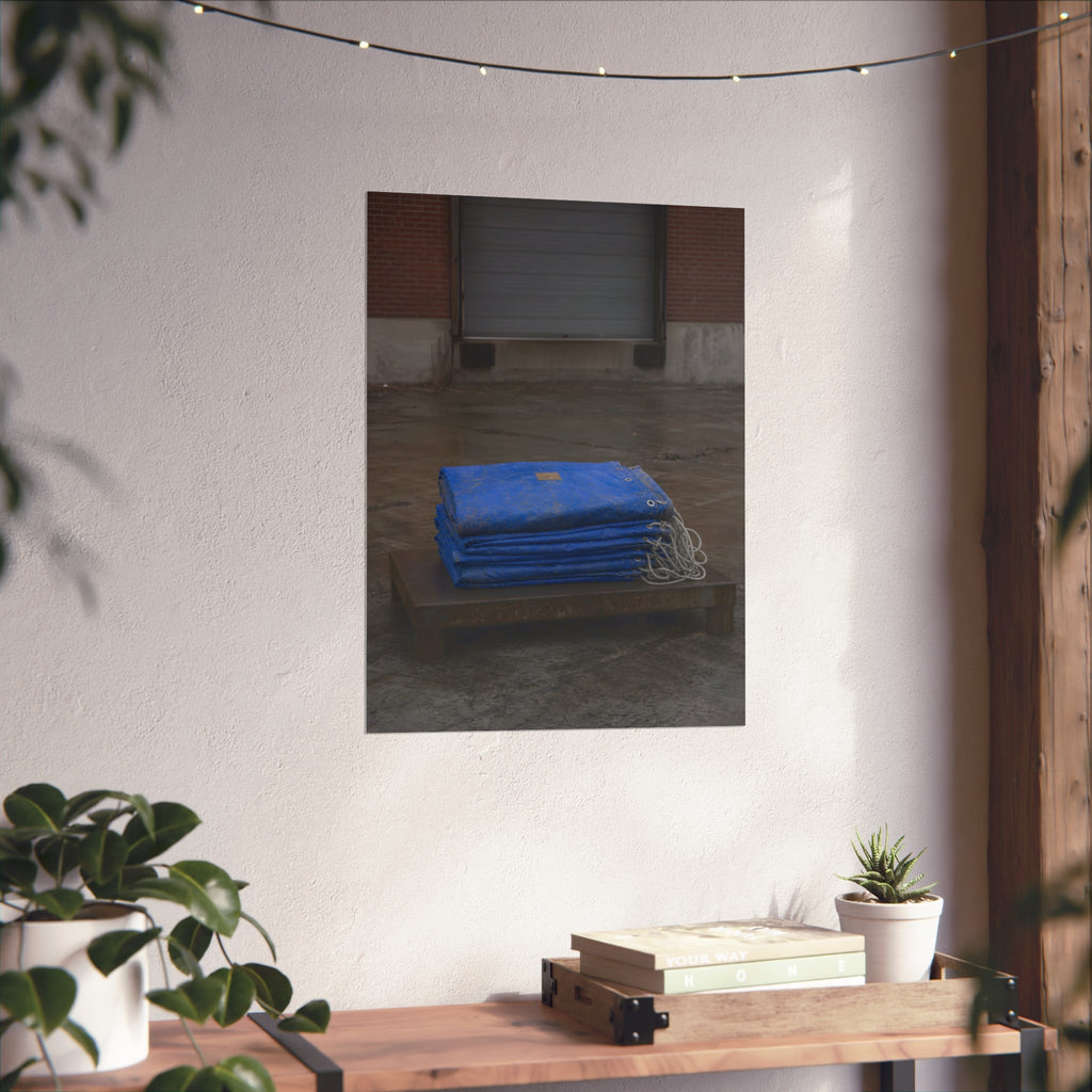 A folded blue tarp stacked on a low wooden platform by a loading dock. The Repaired Fold by Garabaggio, fine art print hanging in a room.