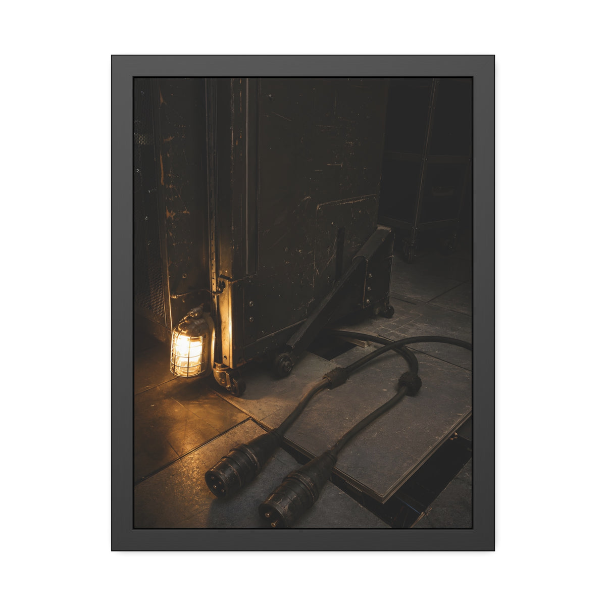 Two thick power cables unplugged on the floor beside a caged work lamp. The Redundant Power by Garabaggio, framed fine art print, 18″ x 24″.