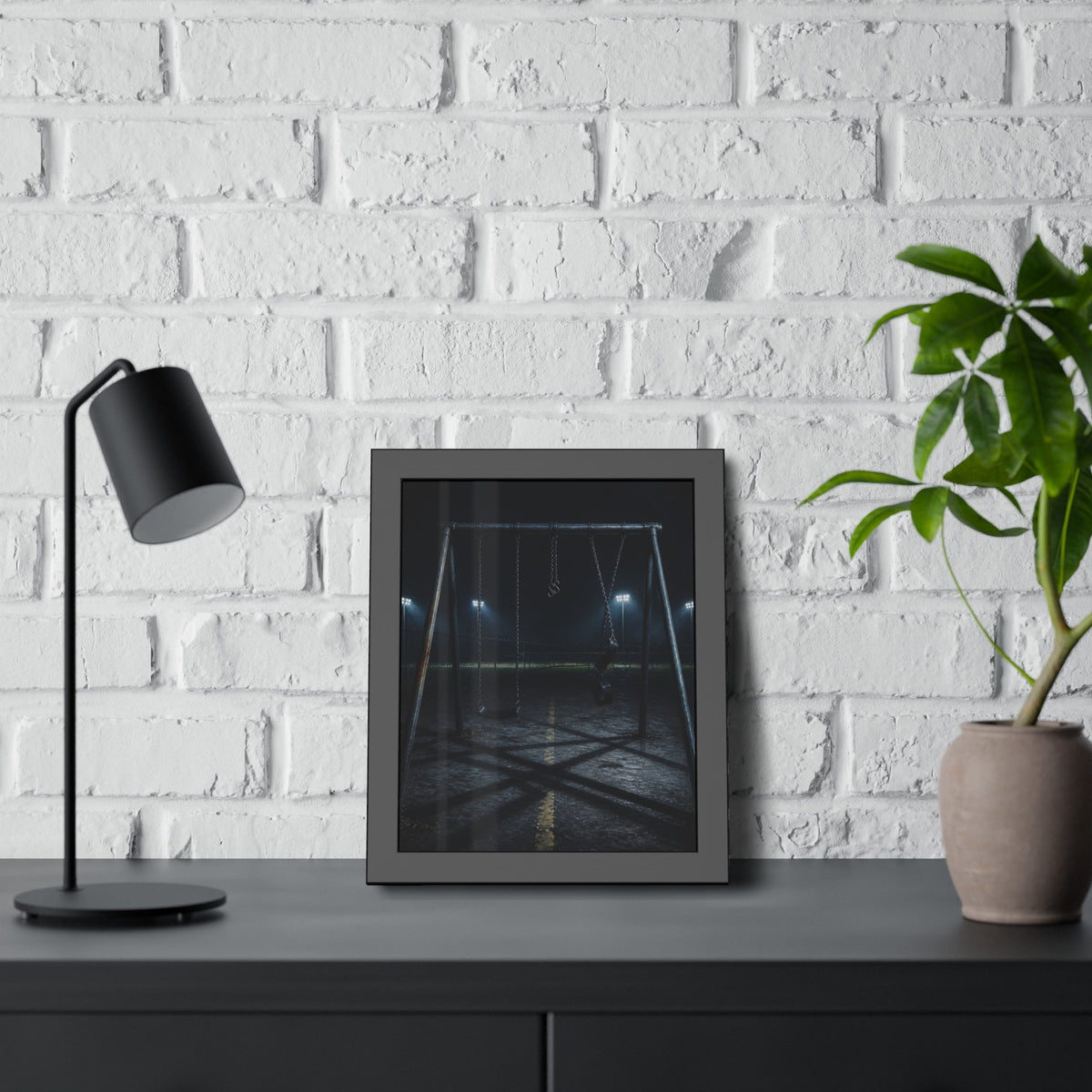 An empty swing set at night with field lights glowing behind it. The Ninth Hour: The Seat Still Moving by Garabaggio, framed fine art print hanging in a room.