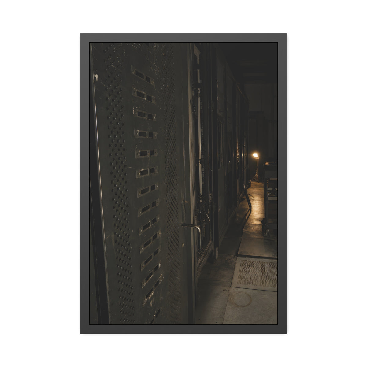 Rows of dark server cabinets with one work lamp glowing far down the aisle. Racks Without Light by Garabaggio, framed fine art print, 24″ x 36″.