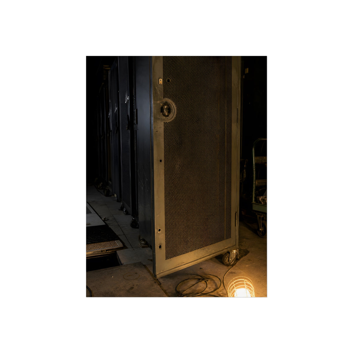 A perforated server rack door in a dark aisle lit by a floor work lamp. The Cold Aisle by Garabaggio, fine art print, 18″ x 24″.