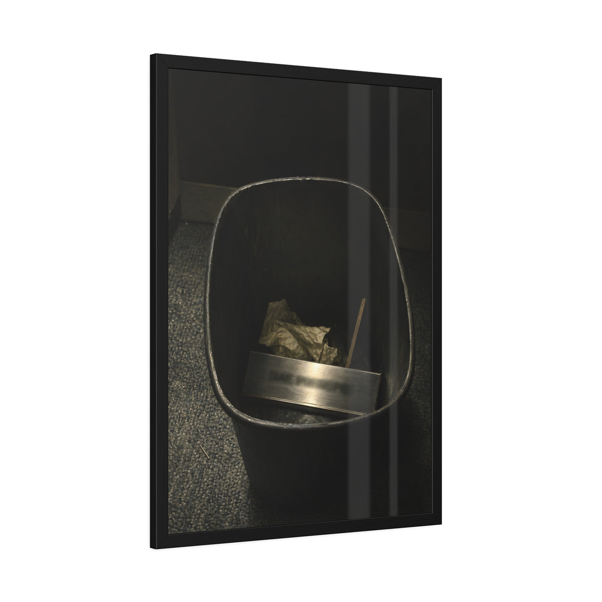 A metal nameplate lying in a black office waste bin on grey carpet. The Nameplate in the Waste Bin by Garabaggio, framed fine art print hanging in a room.