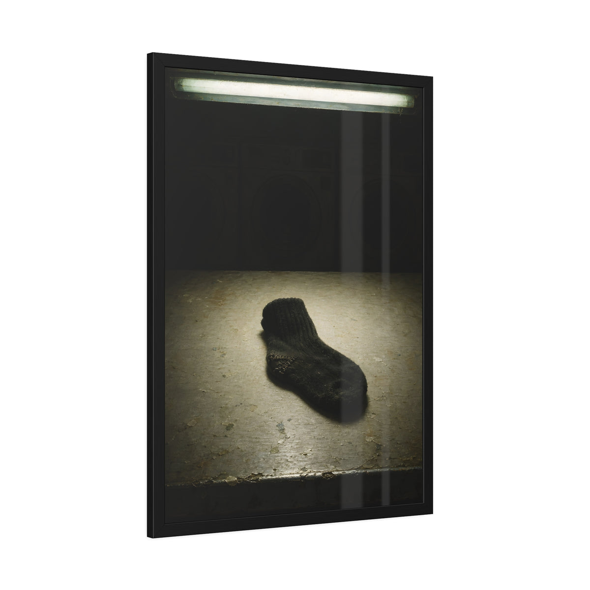 A single dark sock on a laundromat floor before dim washing machines. The Orphaned Sock by Garabaggio, framed fine art print hanging in a room.