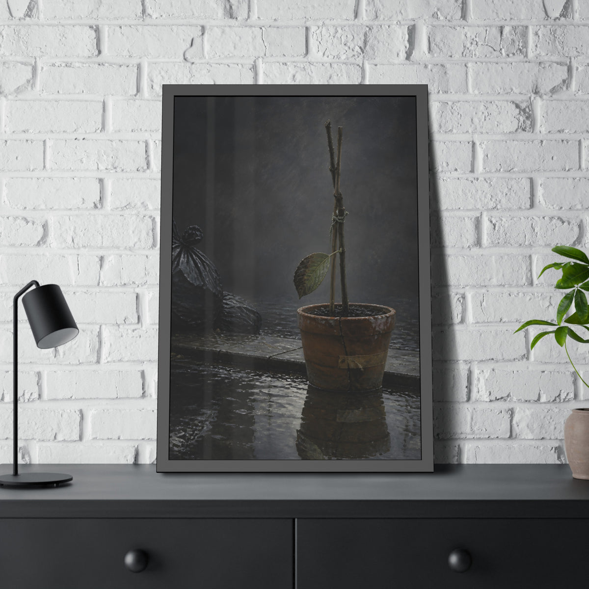 Bare tied stems in a clay pot on flooded pavement, one leaf remaining. The Grey Between by Garabaggio, framed fine art print hanging in a room.