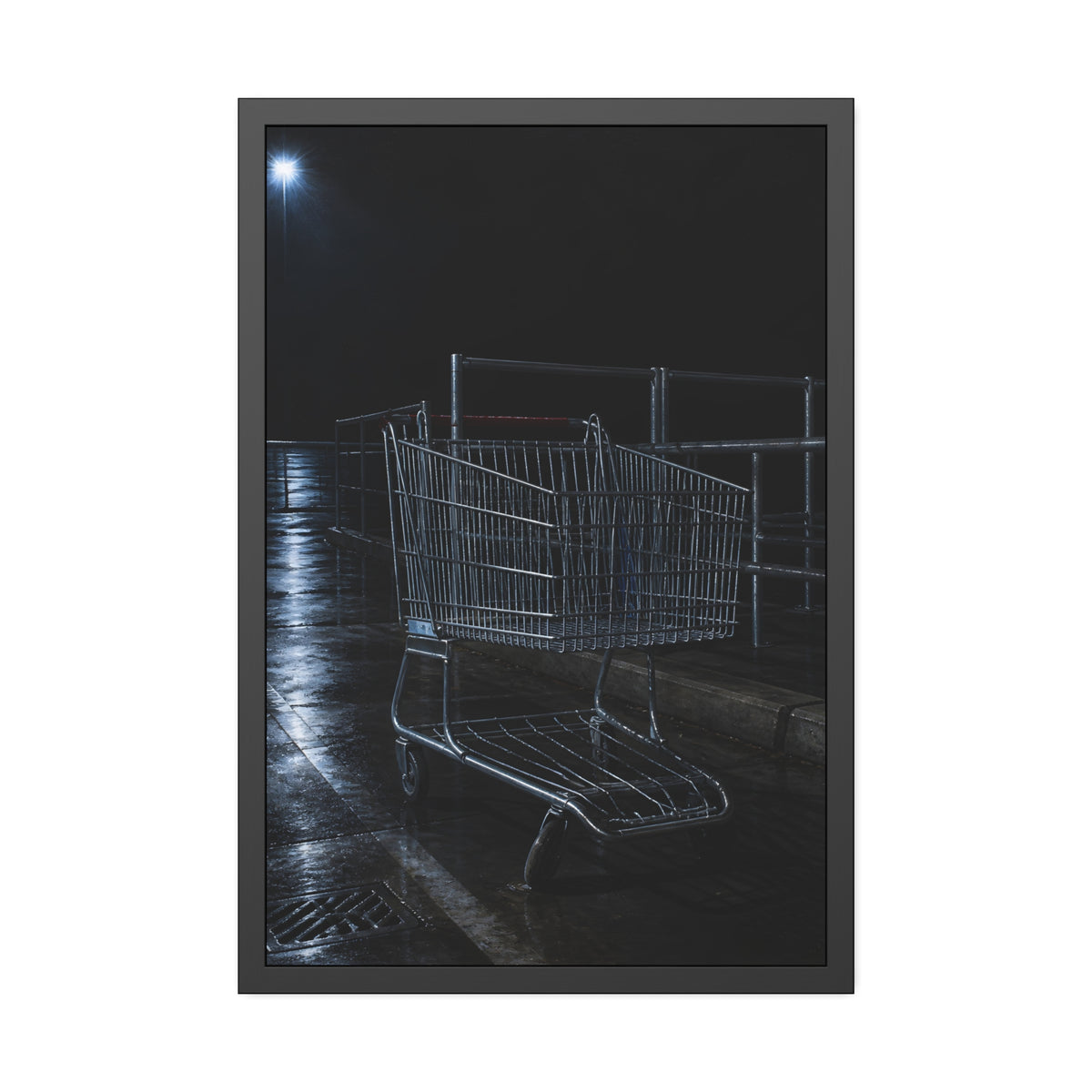 A shopping cart beside its corral on wet asphalt under a lot light. The Cart at the Corral by Garabaggio, framed fine art print, 24″ x 36″.