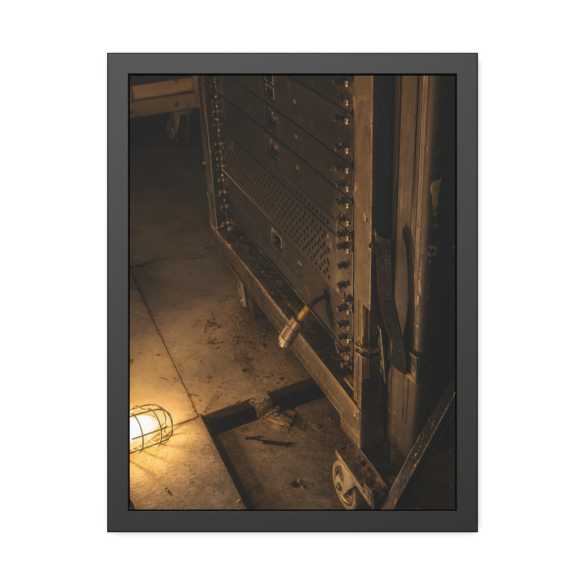 A server rack on a concrete floor beside a caged work light. The Silence at Cutover by Garabaggio, framed fine art print, 18″ x 24″.