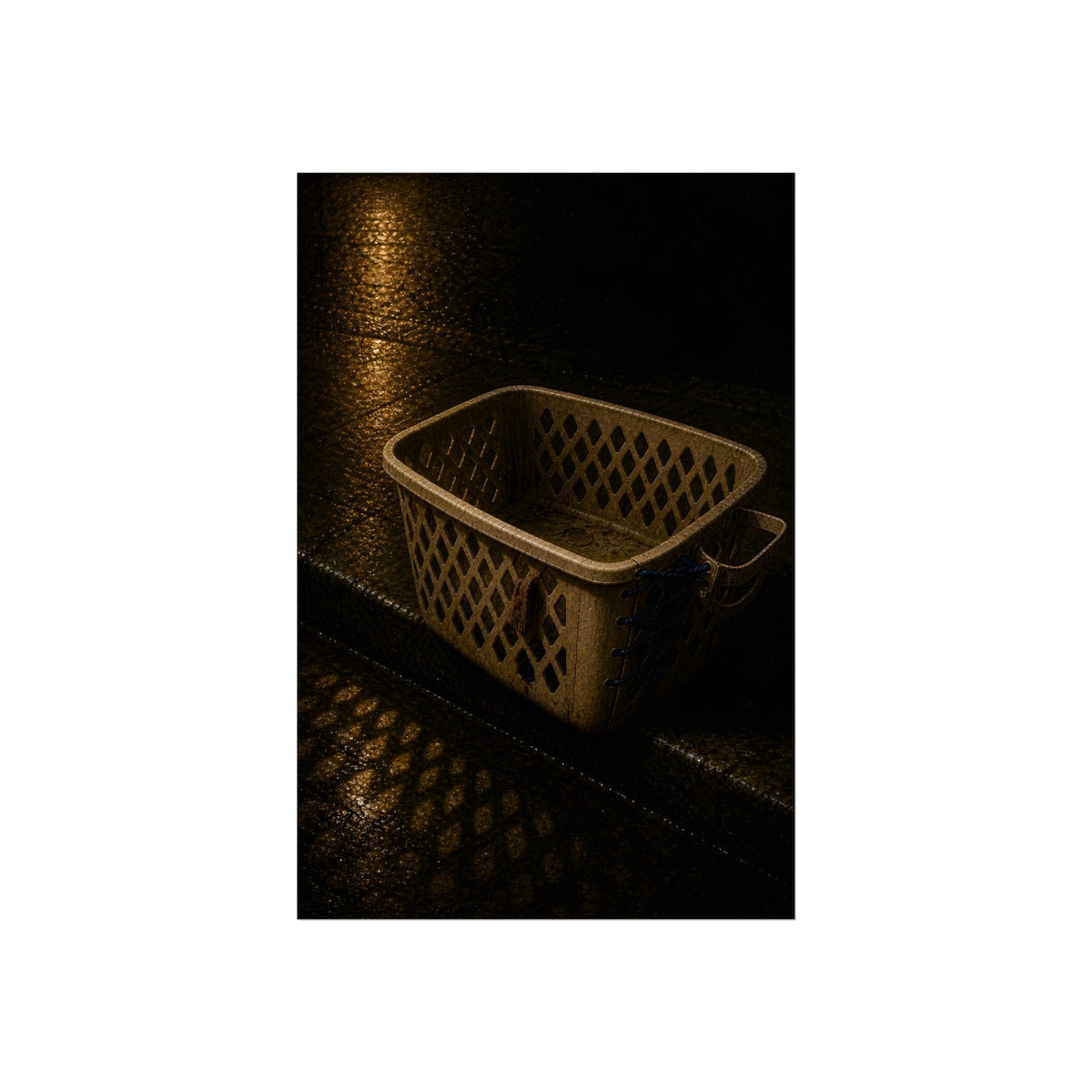 An empty plastic laundry basket on a wet curb under warm street light. The Emptying at the Curb by Garabaggio, fine art print, 24″ x 36″.