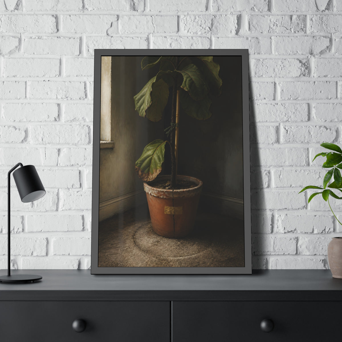A leafy potted fig staked to a cane in a taped clay pot beside a window. The Corner by the Window by Garabaggio, framed fine art print hanging in a room.