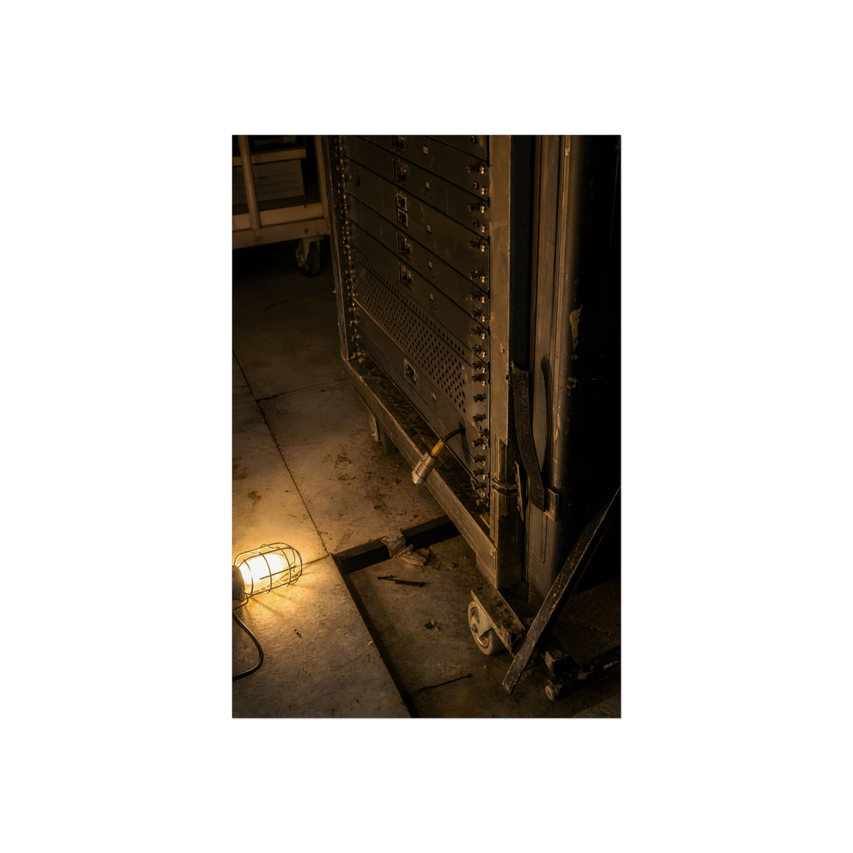 A server rack on a concrete floor beside a caged work light. The Silence at Cutover by Garabaggio, fine art print, 24″ x 36″.